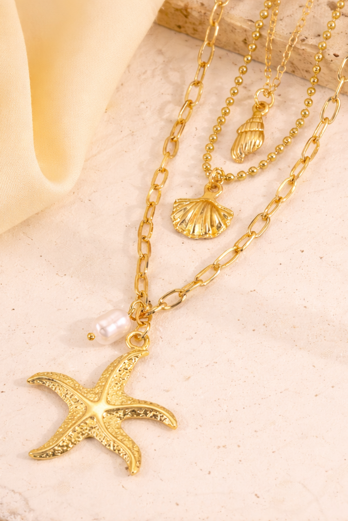 Gold layered stainless steel necklace with starfish, shell, and pearl-inspired charms on beige stone surface